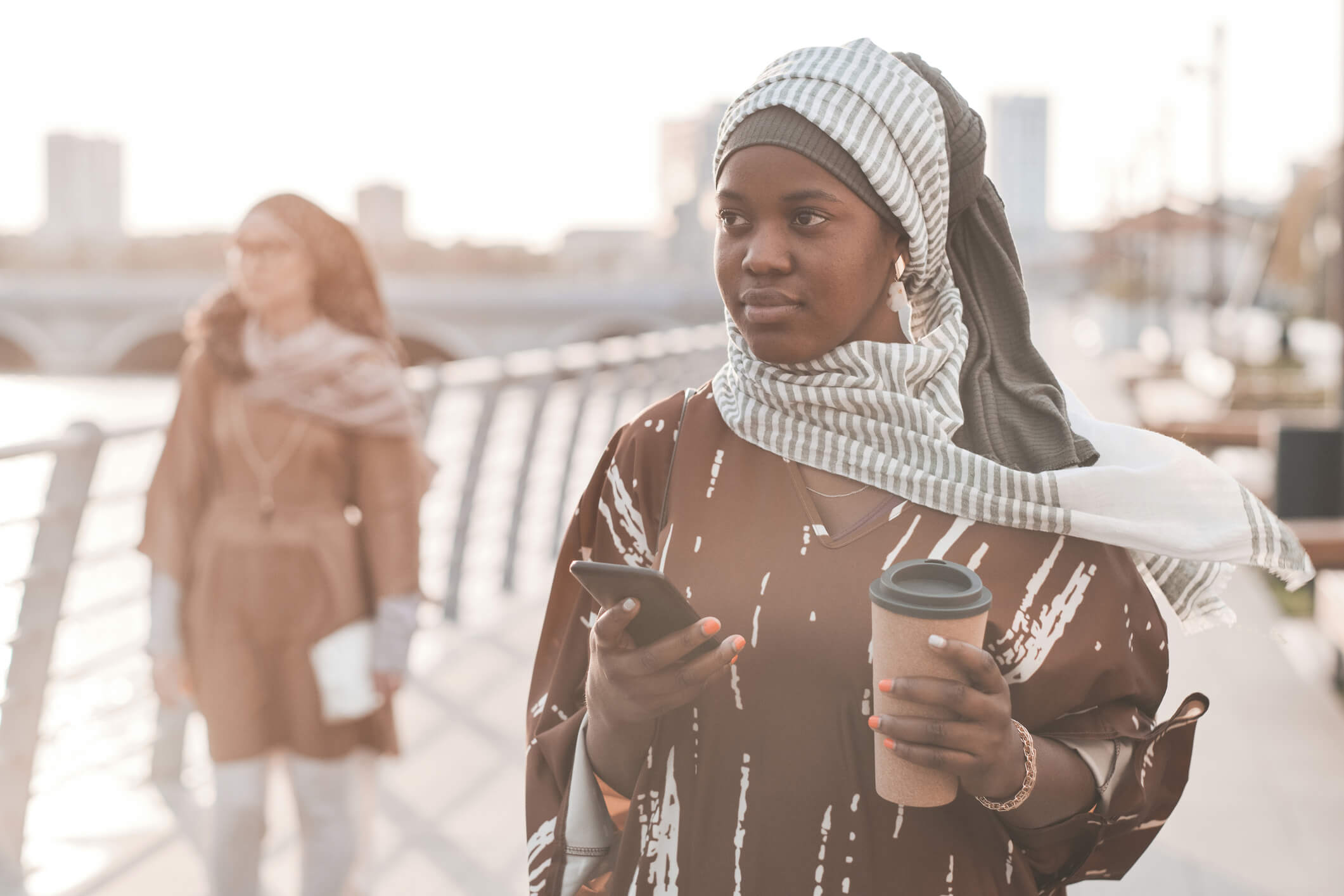 A woman is outside and has a coffee cup in one hand and a cell phone in the other; she has a calm expression.